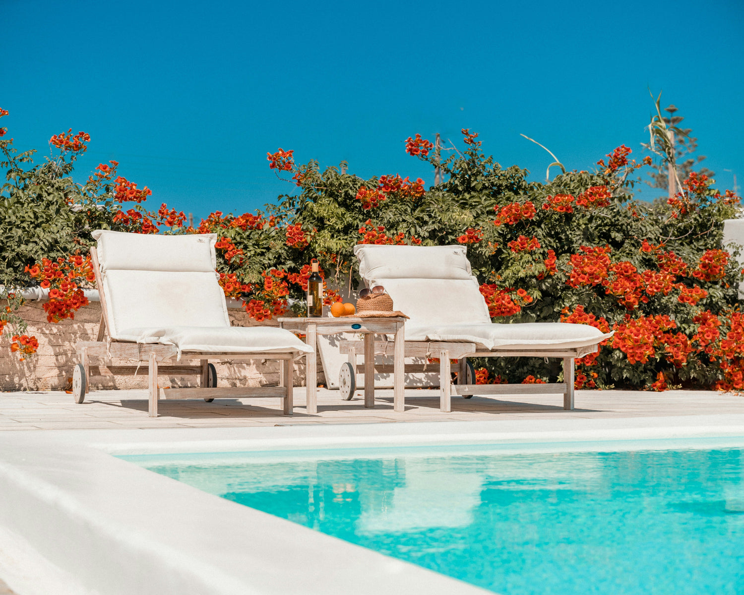 Two white lounge chairs by a swimming pool with red flowers in the background