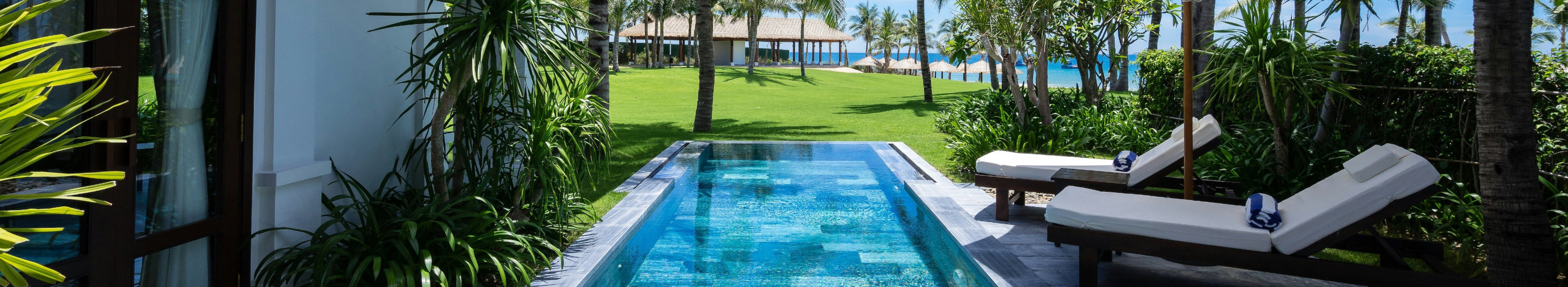 Tropical resort pool area with lounge chairs, palm trees, and clear blue sky.