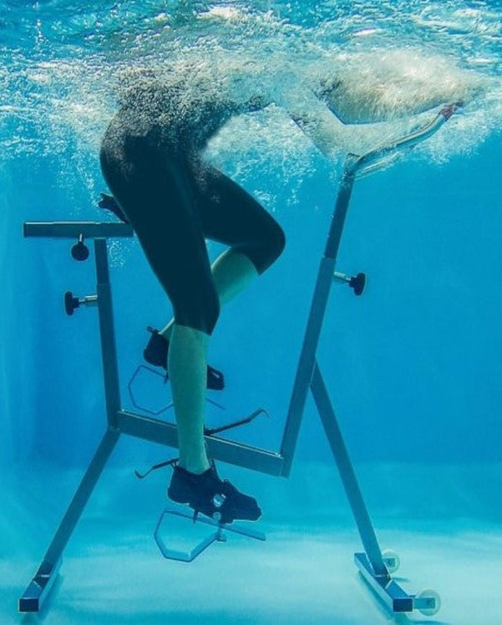 Person using an underwater exercise bike with a shark swimming nearby.