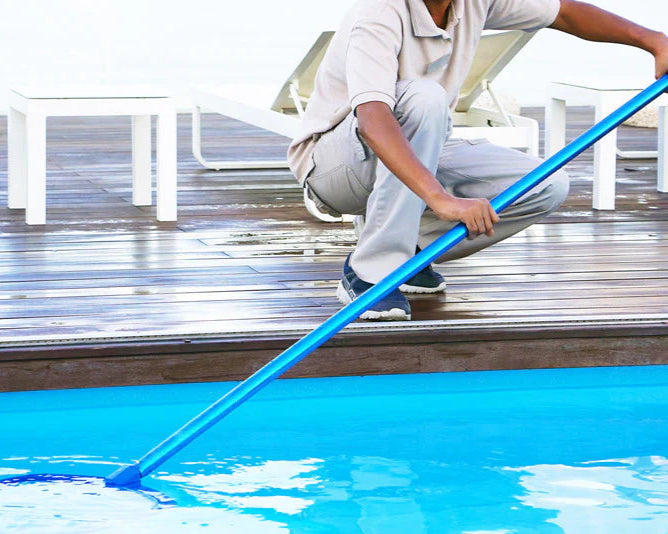 Person cleaning a pool with a blue pool brush on a wooden deck.